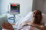 A man with greying stubble lies in a hospital bed with an ECG machine connected to him.