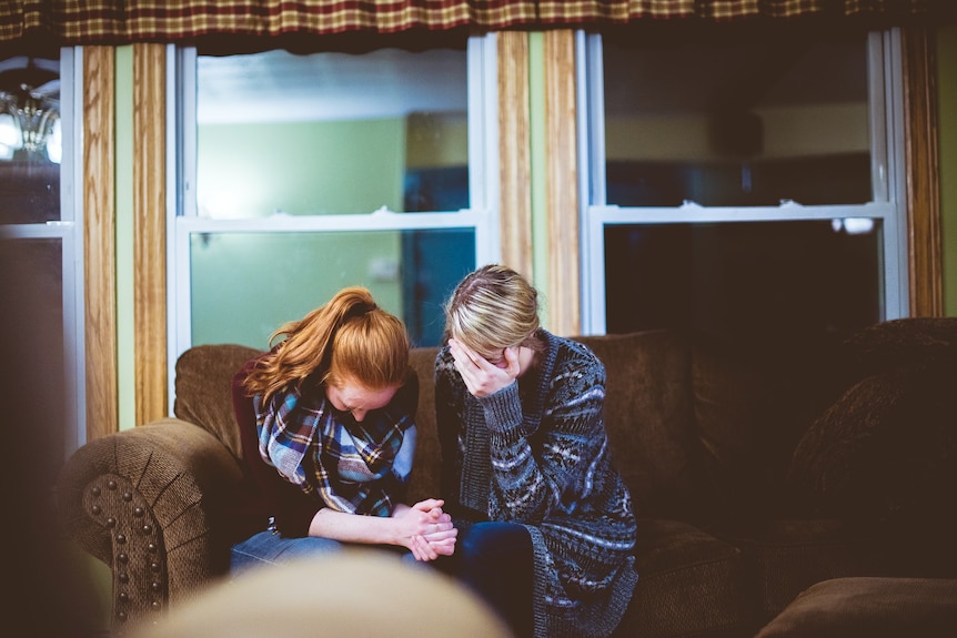 Two women sit on a couch, they appear upset and are gripping each others hands.