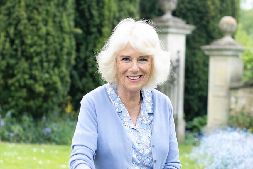 Camilla Parker Bowles sits on a bench in a lush, green garden. She holds a basket of freshly-cut flowers