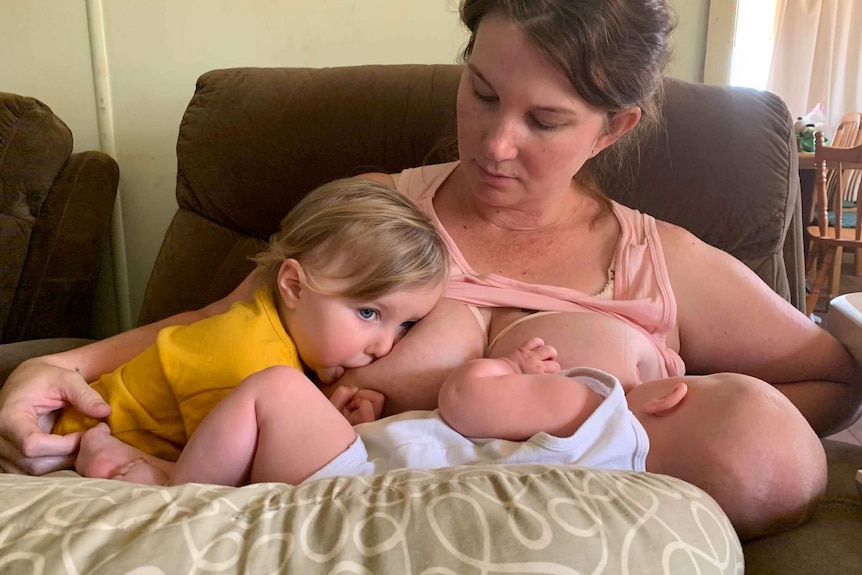 A woman sits on a chair breastfeeding a toddler and newborn baby at the same time.