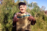 Man in chequered shirt holds old newspaper in front of tree in Esperance backyard
