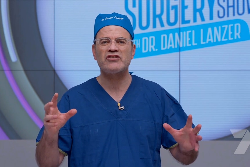 Dr Daniel Lanzer gestures with his hands while talking on the set of a tv show. He is wearing a surgeon's scrubs.