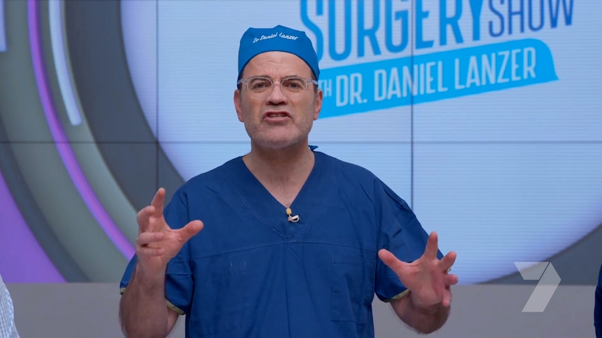 Dr Daniel Lanzer gestures with his hands while speaking on the set of a TV show.  He wears surgeon's gowns.
