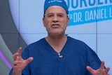 Dr Daniel Lanzer gestures with his hands while talking on the set of a tv show. He is wearing a surgeon's scrubs.