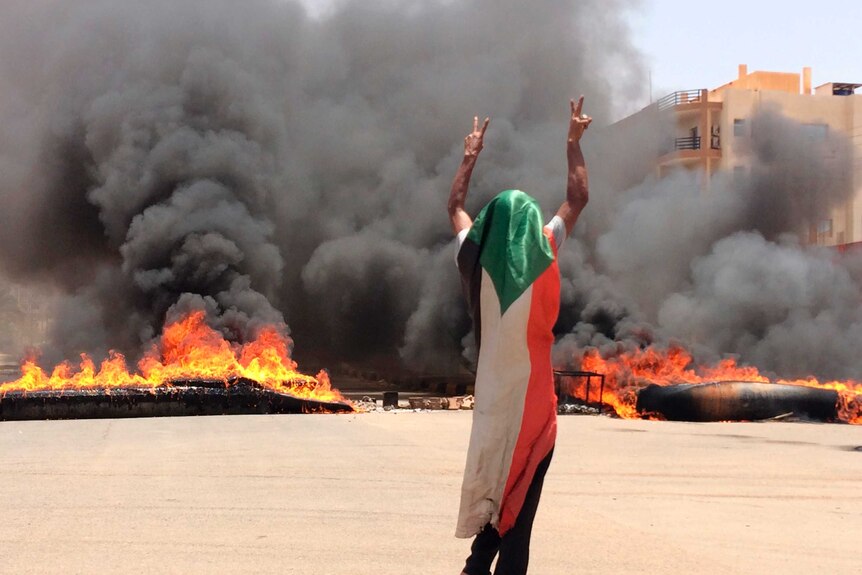 A man draped in a flag holds up the victory sign as fire and black smoke billow in the background.