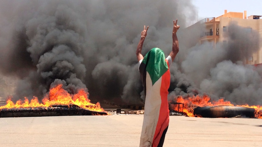 A man draped in a flag holds up the victory sign as fire and black smoke billow in the background.