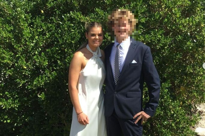 A young woman wearing a long white dress standing with a young man wearing a suit (his face has been pixellated)