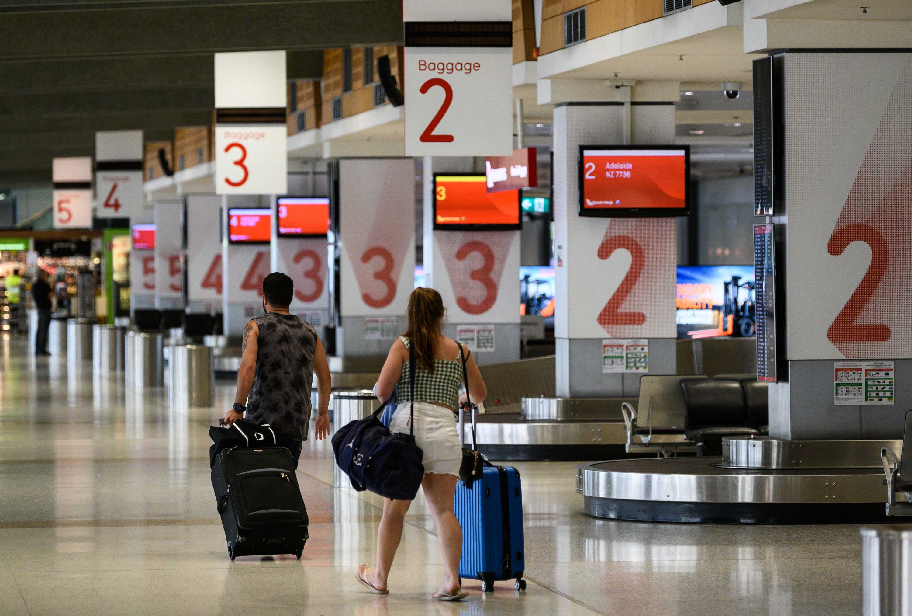 A young man and woman walk away with suitcases in from of empty baggage arrivals area.