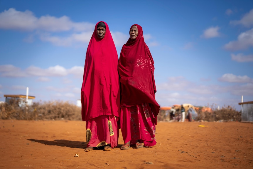 two women in red clothes stand on red dirt outside a camp