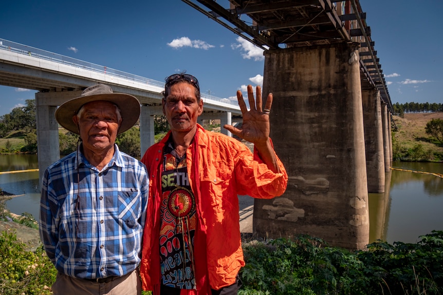 Two men stand under an old bridge across a river in a rural setting, with another newer bridge behind them.