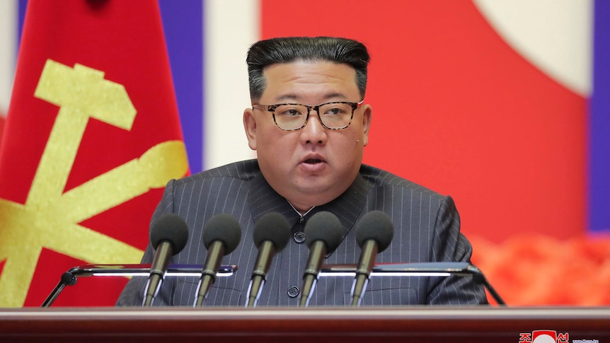 A heavy Korean man wearing a striped suit and horn-rimmed glasses speaks into microphones in front of the North Korean flag.