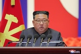 A heavy Korean man wearing a striped suit and horn-rimmed glasses speaks into microphones in front of the North Korean flag.