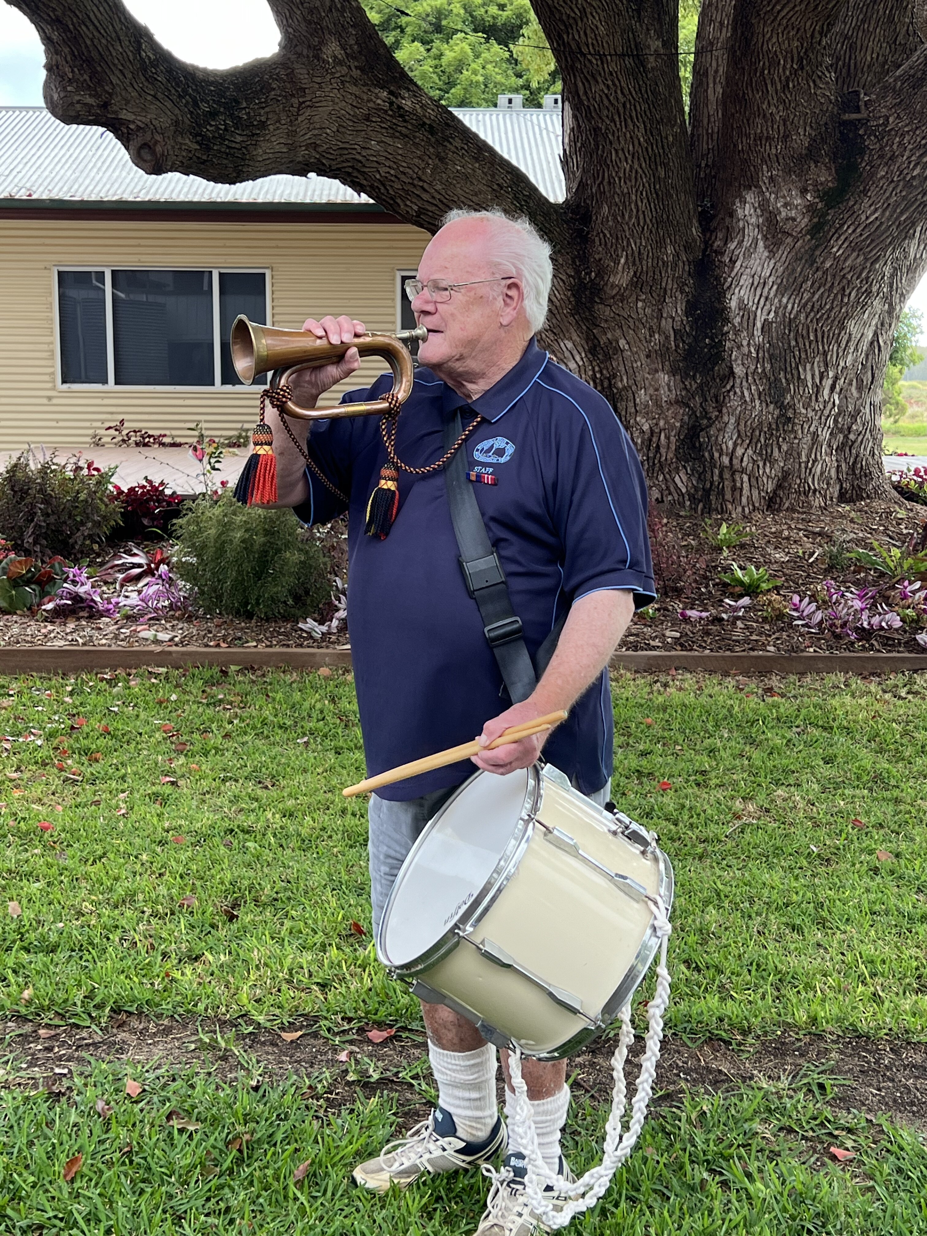 Man in blue polo plays the bugle as he carries a drum strapped over his shoulder.