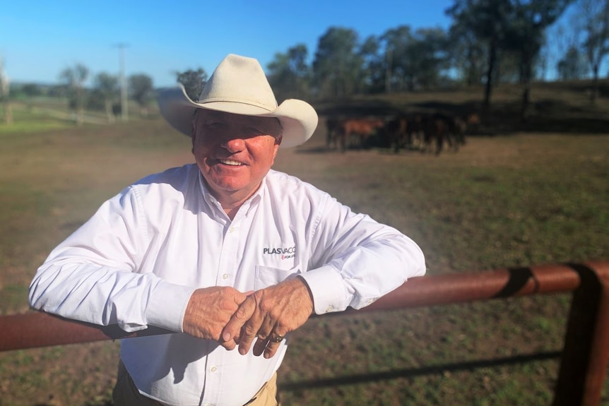 A man in a white shirt and hat stands in a paddock with horses behind him.