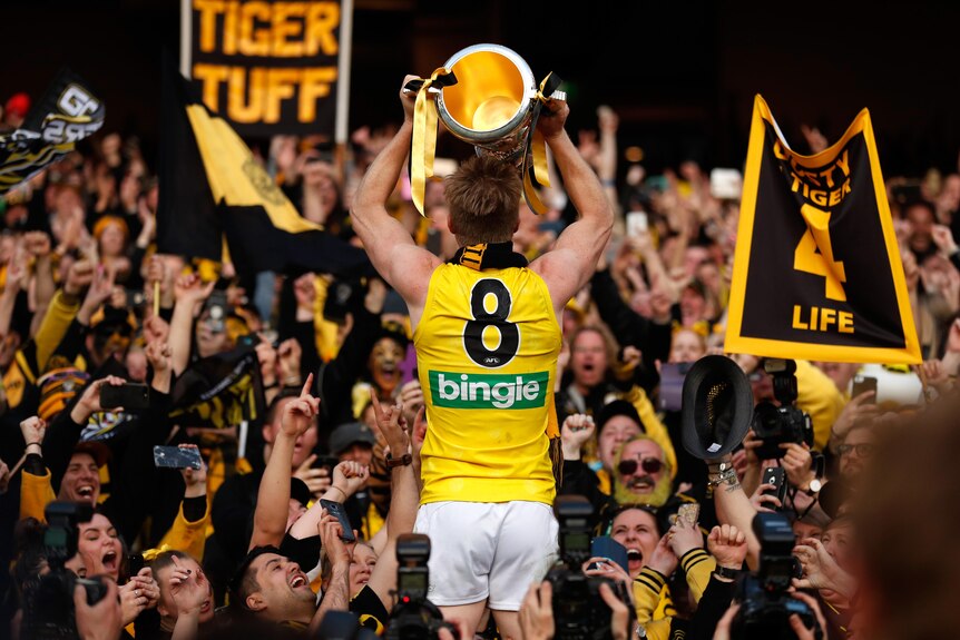 A Richmond AFL star stands with his back to camera holding the premiership cup up, surrounded by fans.