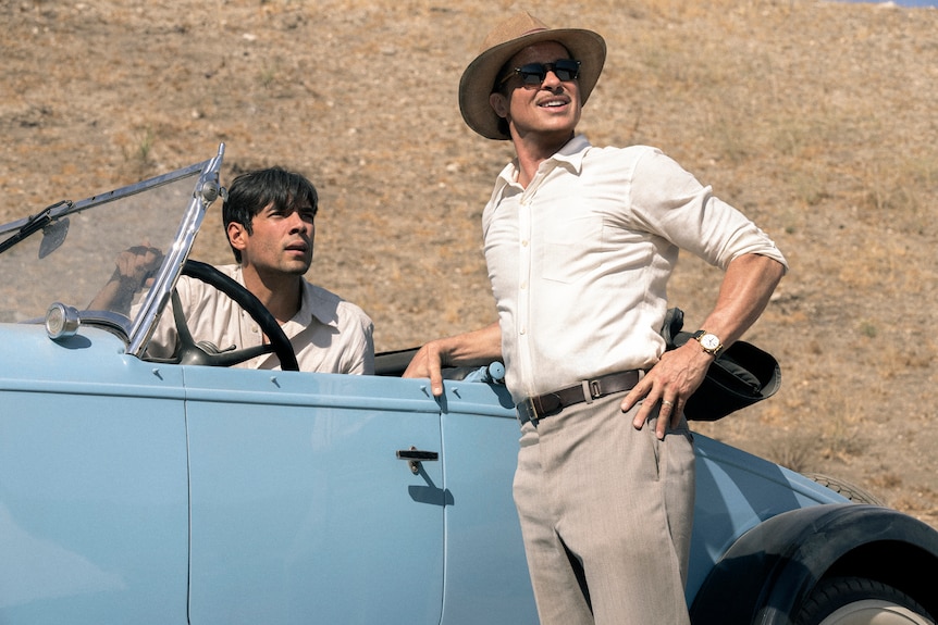 On a bright day, a Hispanic man sits in a blue retro car, another man leaning against it, both looking in same direction.