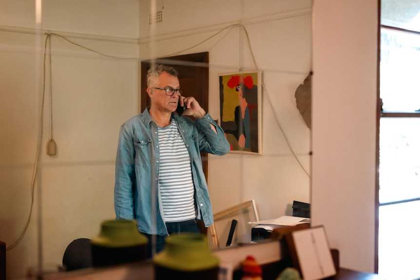 A man wearing glasses standing inside with phone to his ear is seen in the reflection of a mirror.