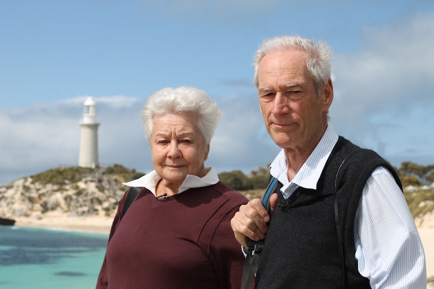 An older woman and man stand in front of a bay, a lighthouse on the far headland.