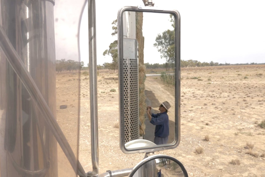 Looking at the side mirror of a truck seeing the reflection of a man adjusts straps on the back of a truck carrying a load