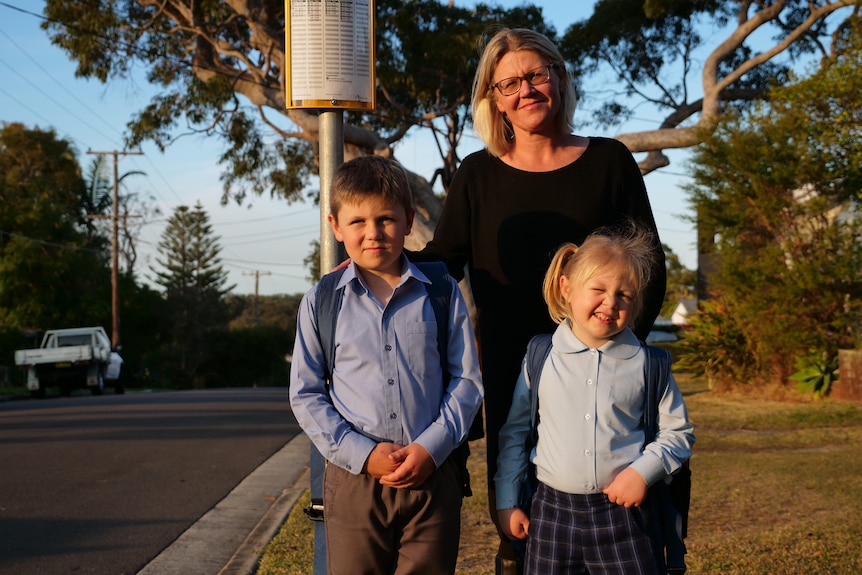 A woman stands behind two children in school uniform on a suburban street.