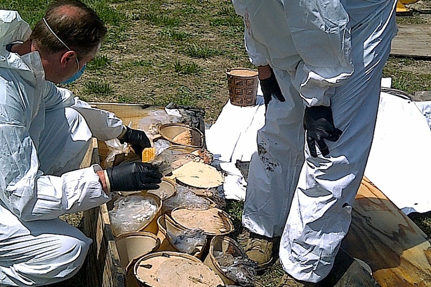 Two men in protective outfits look over substances in buckets near a burial site.