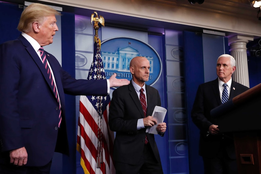 President Donald Trump gestures to Vice President Mike Pence as Dr Stephen Hahn as they stand behind a podium.