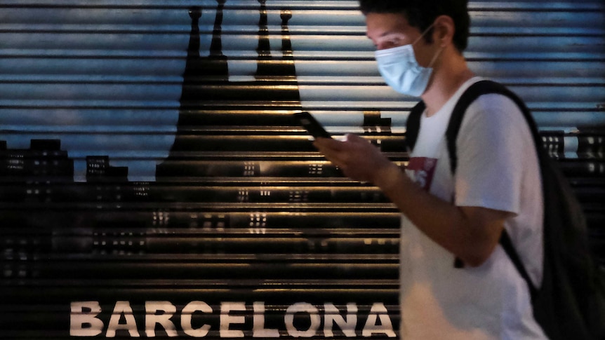 A young man wearing a face mask looks at his phone as he walks past a Barcelona sign on garage door.