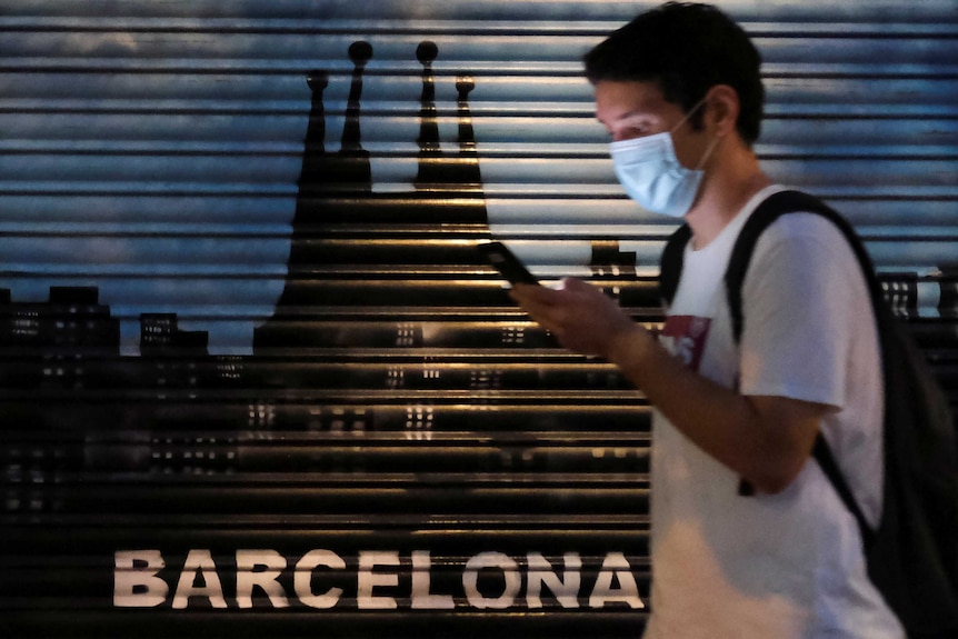 A young man wearing a face mask looks at his phone as he walks past a Barcelona sign on garage door.