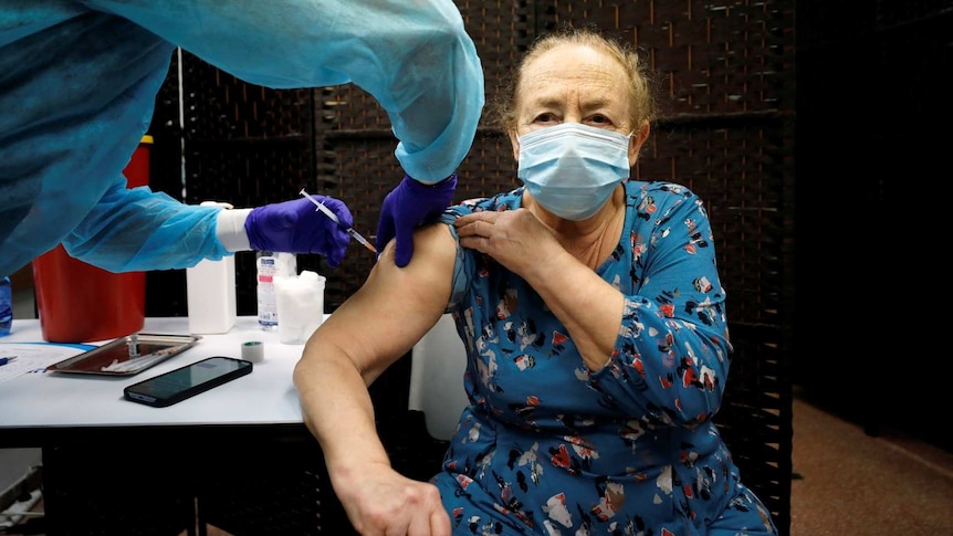 a woman in a blue dress and a mask receives a vaccination from a person in PPE