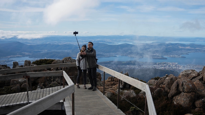 A couple stand on a boardwalk taking a pic with a selfie stick with Hobart landscape behind 