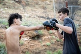 A teenager with shirt off throws a punch with gloves on, and hits pads held by another teenager with shirt on 