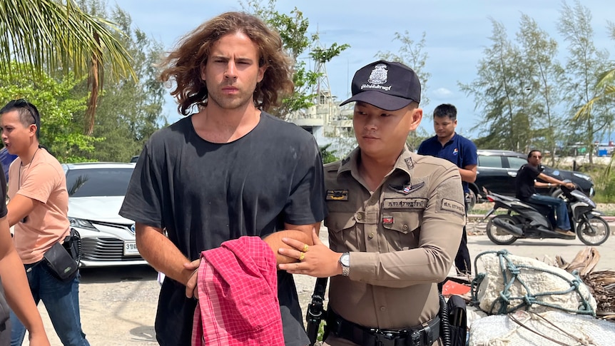A man with long hair in handcuffs is escorted by a police officer