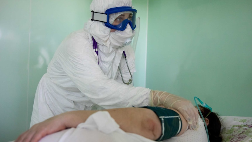 Health worker in white suit with blue goggles places hand on a patient
