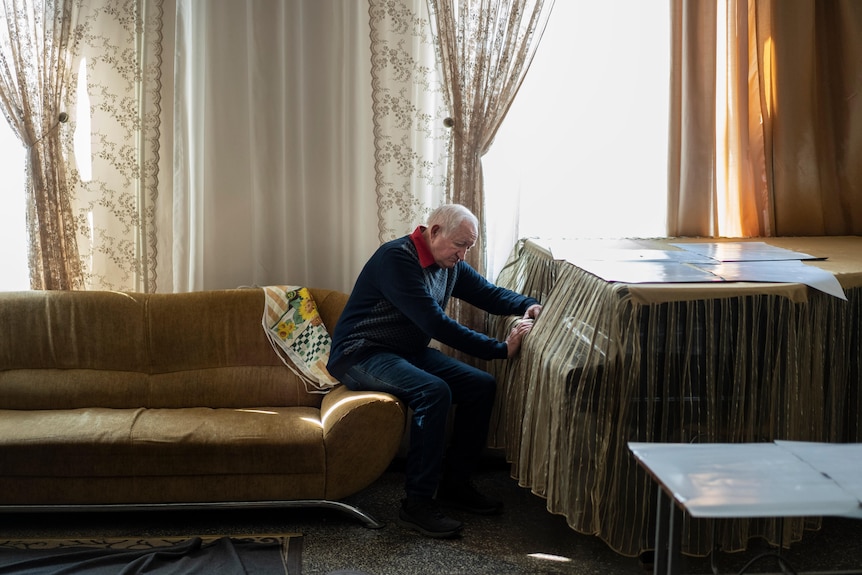 An elderly white-haired man sits in his house alone with a distressed look on his face.
