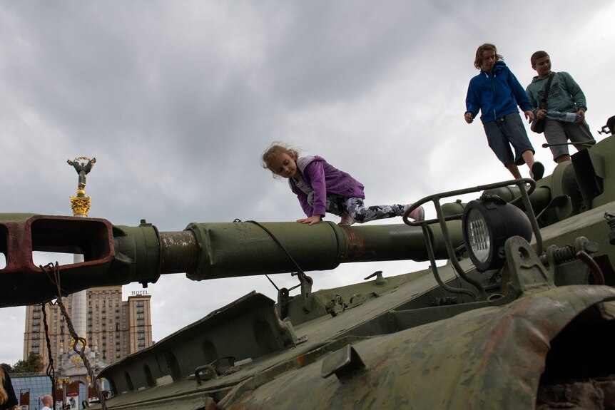 Children climb over a disused tank, a statue is visible in the background against a grey sky