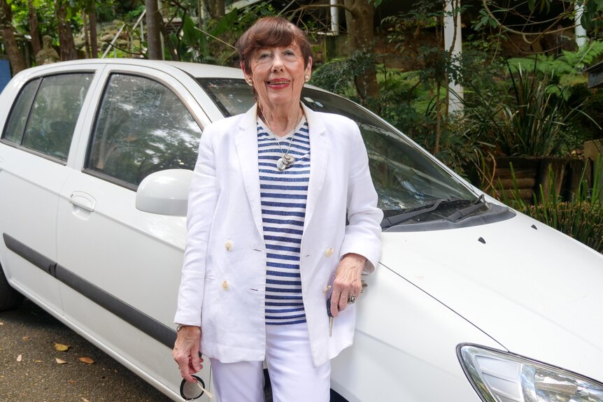 An elderly woman in a white suit stands in front of a small white car. 
