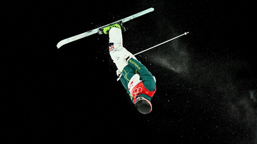 Jakara skis through the air upside down against the night sky.