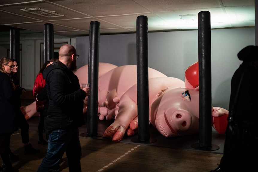 Interior, hall. 3-metre long inflatable pink cartoonish pig behind floor-to-ceiling black inflatable bars. Audience walk by.