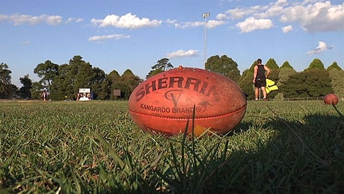 A red AFL football lies on grass at a country oval, players in the background.