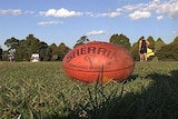 A red AFL football lies on grass at a country oval, players in the background.
