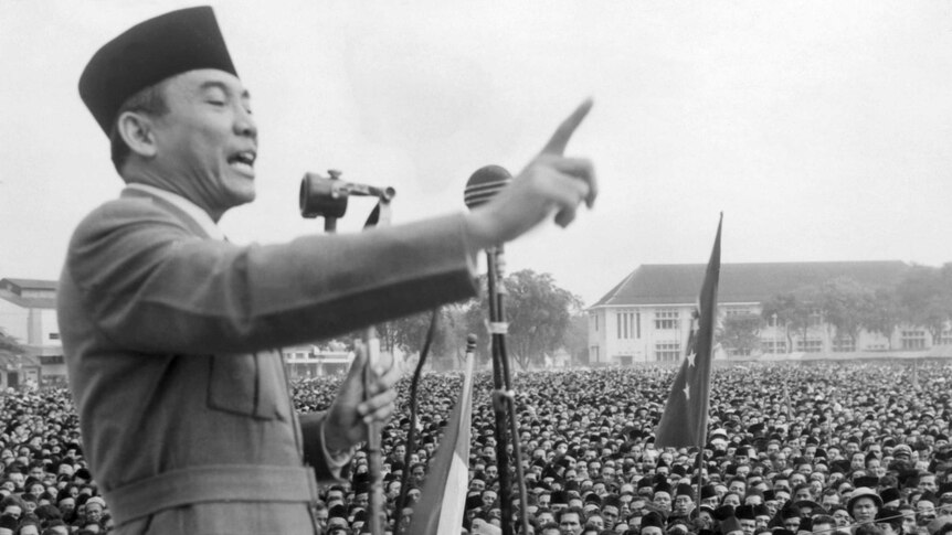 A black and white image shows a man addressing a large crowd, holding his finger in the air.