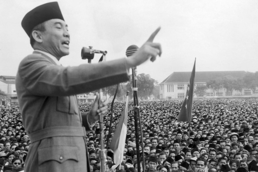 A black and white image shows a man addressing a large crowd, holding his finger in the air.