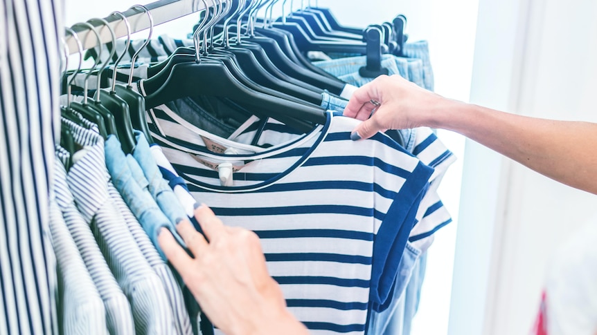 a shopper browsing through a rack of clothes stops to look closely at a t-shirt