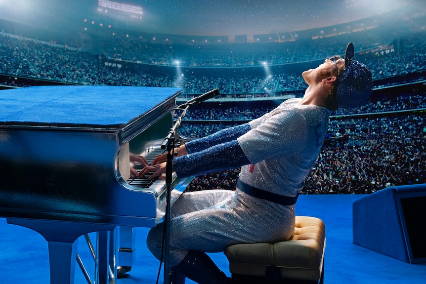 A man in a glittering suit plays a piano on stage