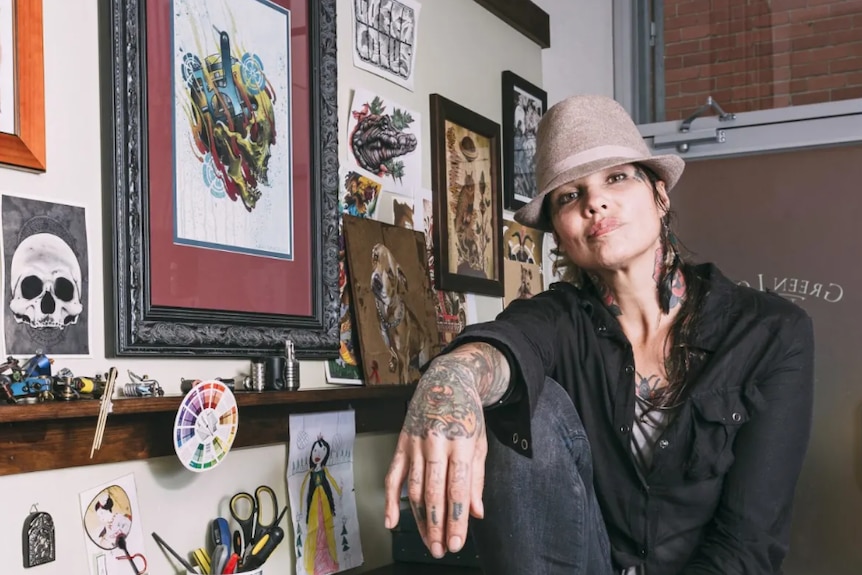 A heavily tattooed young woman in a trendy hat poses in a tattoo parlour.