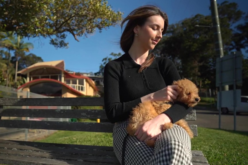 A woman sits on a park bench with a small dog on her lap.