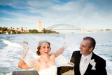 A laughing bride and groom on a boat in Sydney Harbour with the bridge behind them. She is trying to catch her veil in the wind.