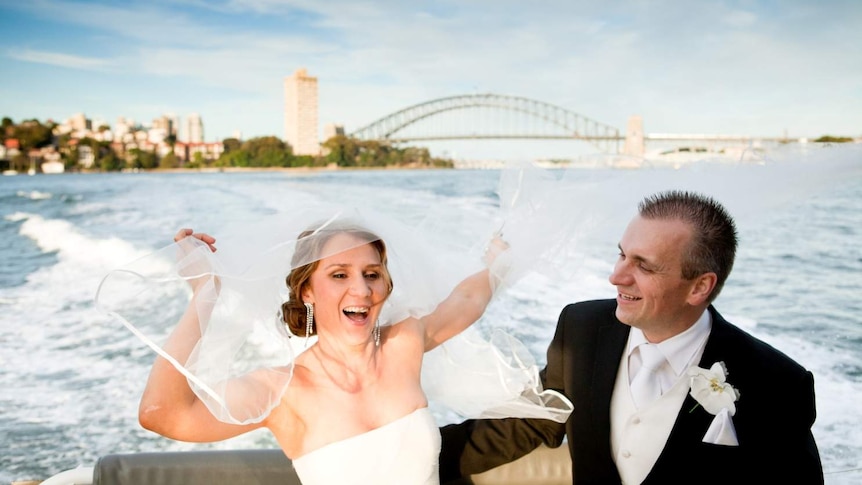 A laughing bride and groom on a boat in Sydney Harbour with the bridge behind them. She is trying to catch her veil in the wind.