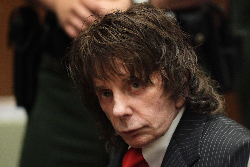 A man with shaggy hair looks towards a camera in a close up shot taken while he is sitting inside a courtroom.