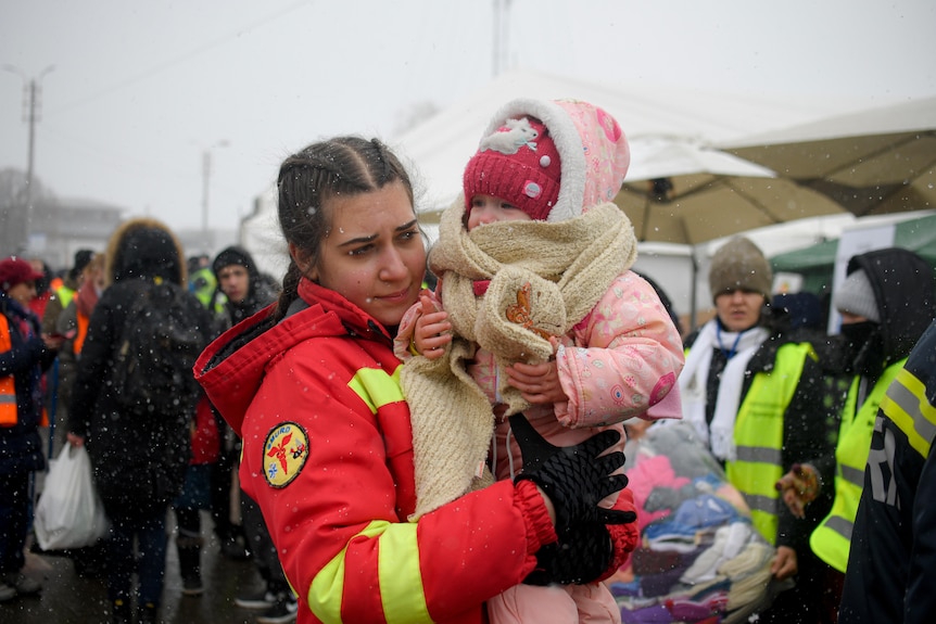 A woman with braided hair wearing an orange jacket carries a small crying child in her arms.